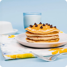 Stack of pancakes with chocolate chips on a plate with a glass of milk against a light blue background.