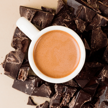 White mug filled with a brown liquid surrounded by dark chocolate pieces on a light background